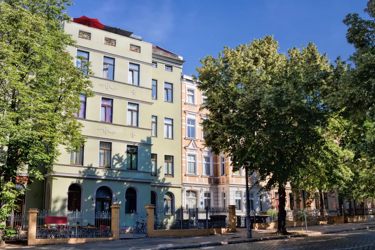 Historisches Mehrfamilienhaus mit grüner Fassade und Bäumen an einer städtischen Straße.